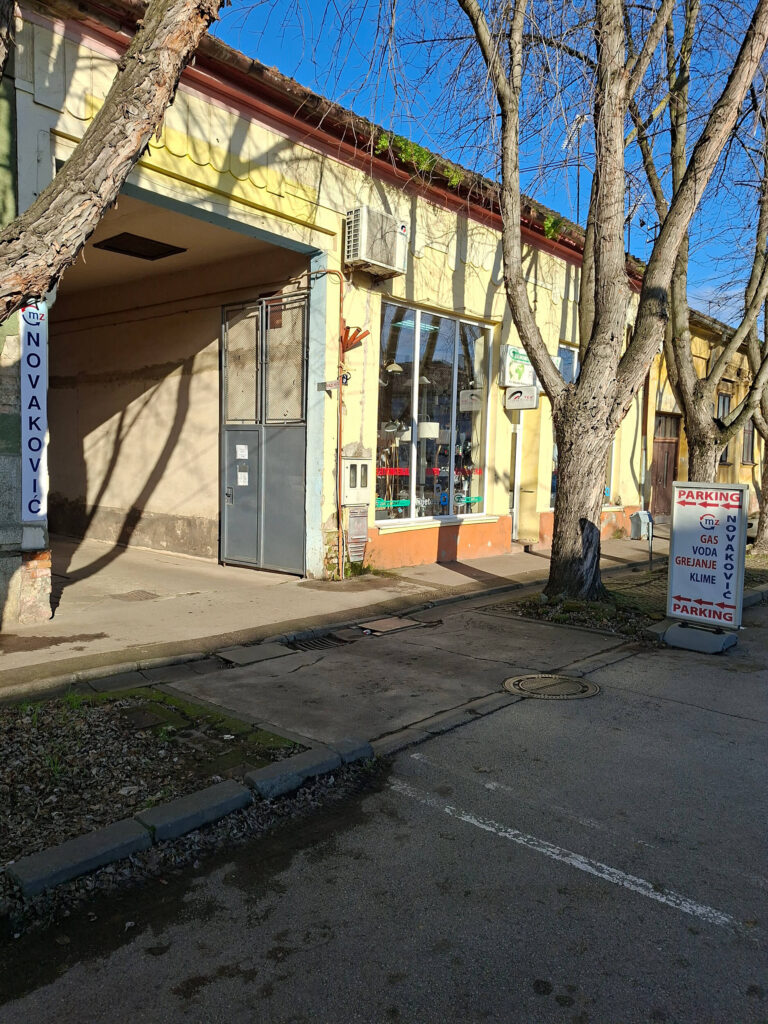 Prilaz i ulaz u dvorište prodavnice M.Z. Novaković u Kikindi sa putokazom za besplatan parking za kupce. Ulaz kroz kapiju pored reklame za grejanje, vodovod, belu tehniku i kupatilo.