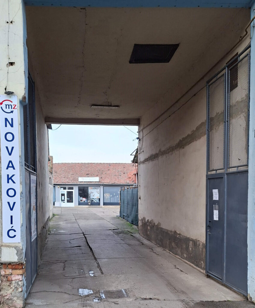 Prolaz u dvorište prodavnice M.Z. Novaković u Kikindi koji vodi do besplatnog parkinga i glavnog ulaza prodavnice. Reklama za grejanje, vodovod, belu tehniku i kupatilo vidljiva na ulaznoj kapiji.